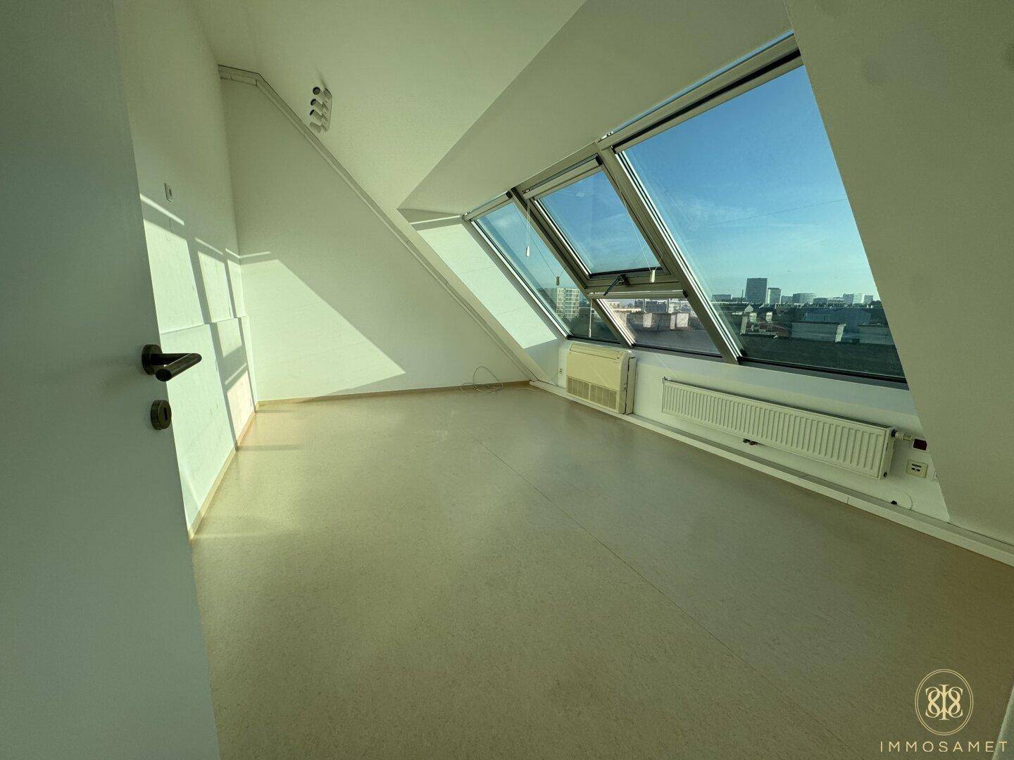 Leerer Dachgeschossraum mit schrägen Wänden und großen Fenstern, die viel Sonnenlicht hereinlassen.