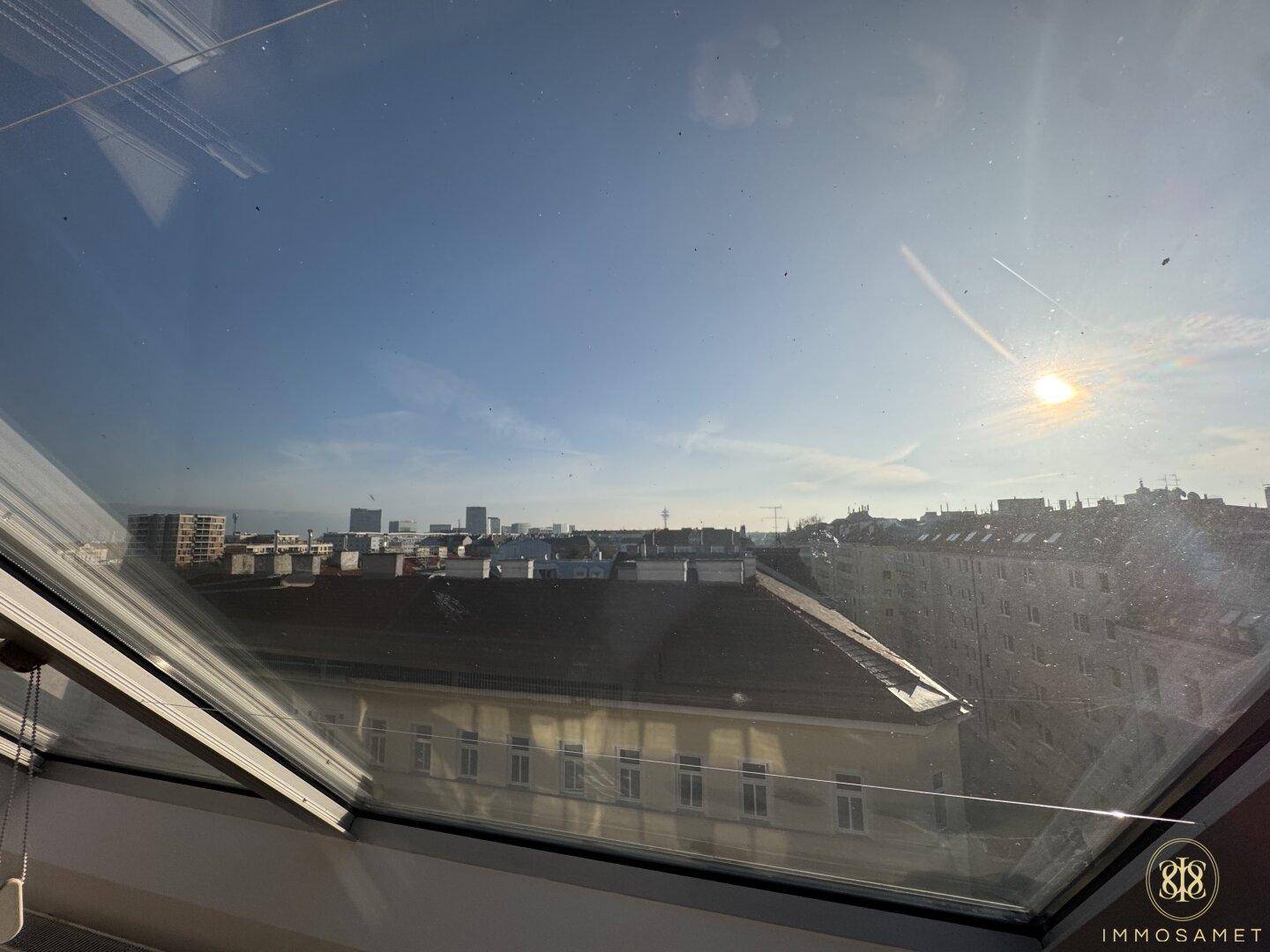 Weitblick über die Stadt aus einem Dachfenster, mit Gebäuden und blauem Himmel.