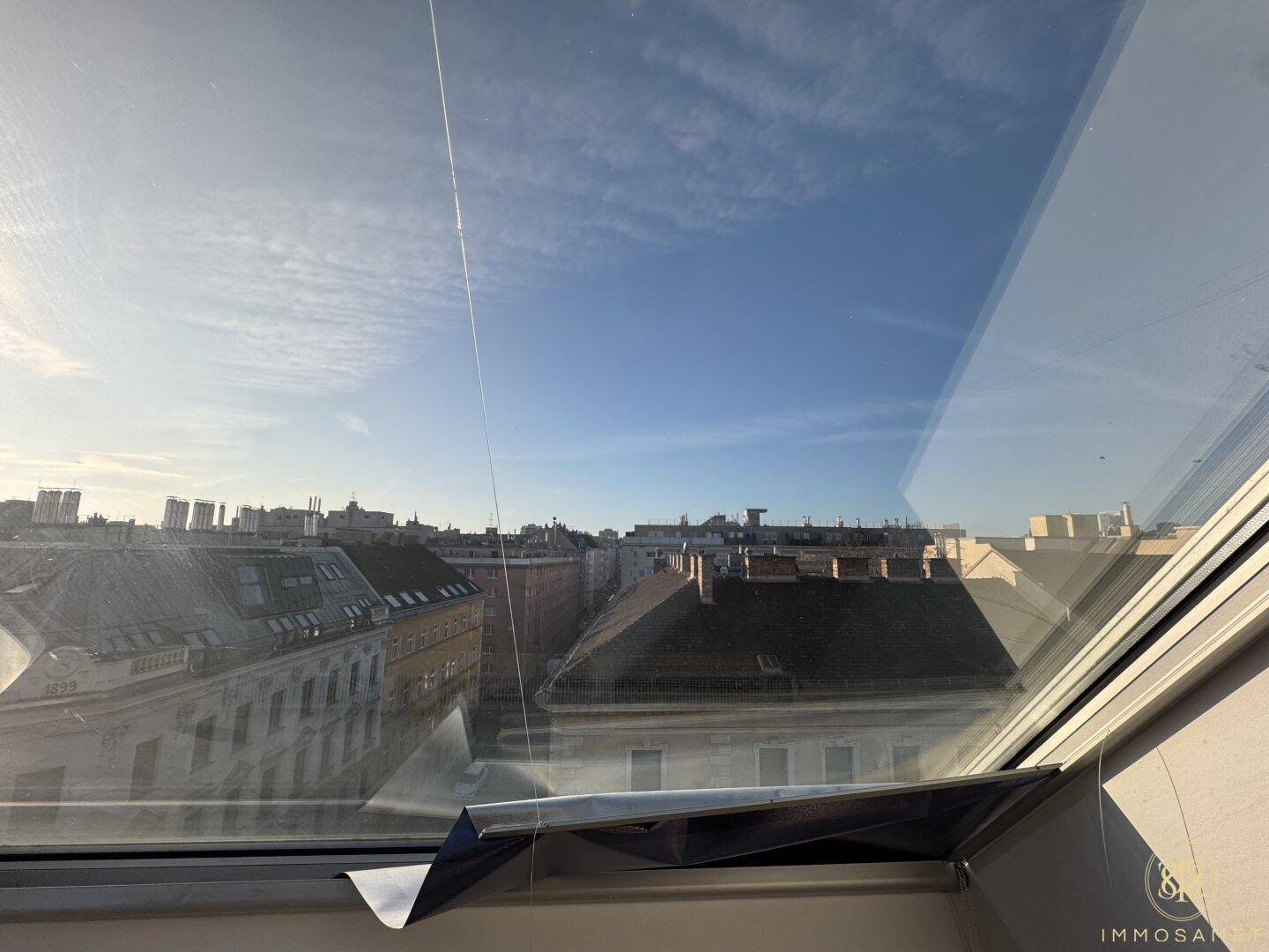 Weitblick über die Stadt aus einem Dachfenster, mit Gebäuden und blauem Himmel.