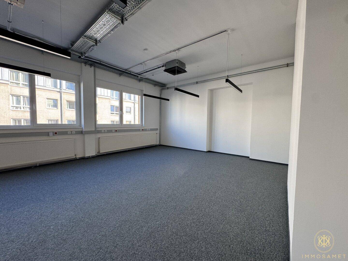Heller, weitläufiger Bürobereich mit großen Fenstern, grauem Teppichboden und sichtbaren Deckeninstallationen, bietet vielseitige Gestaltungsmöglichkeiten.