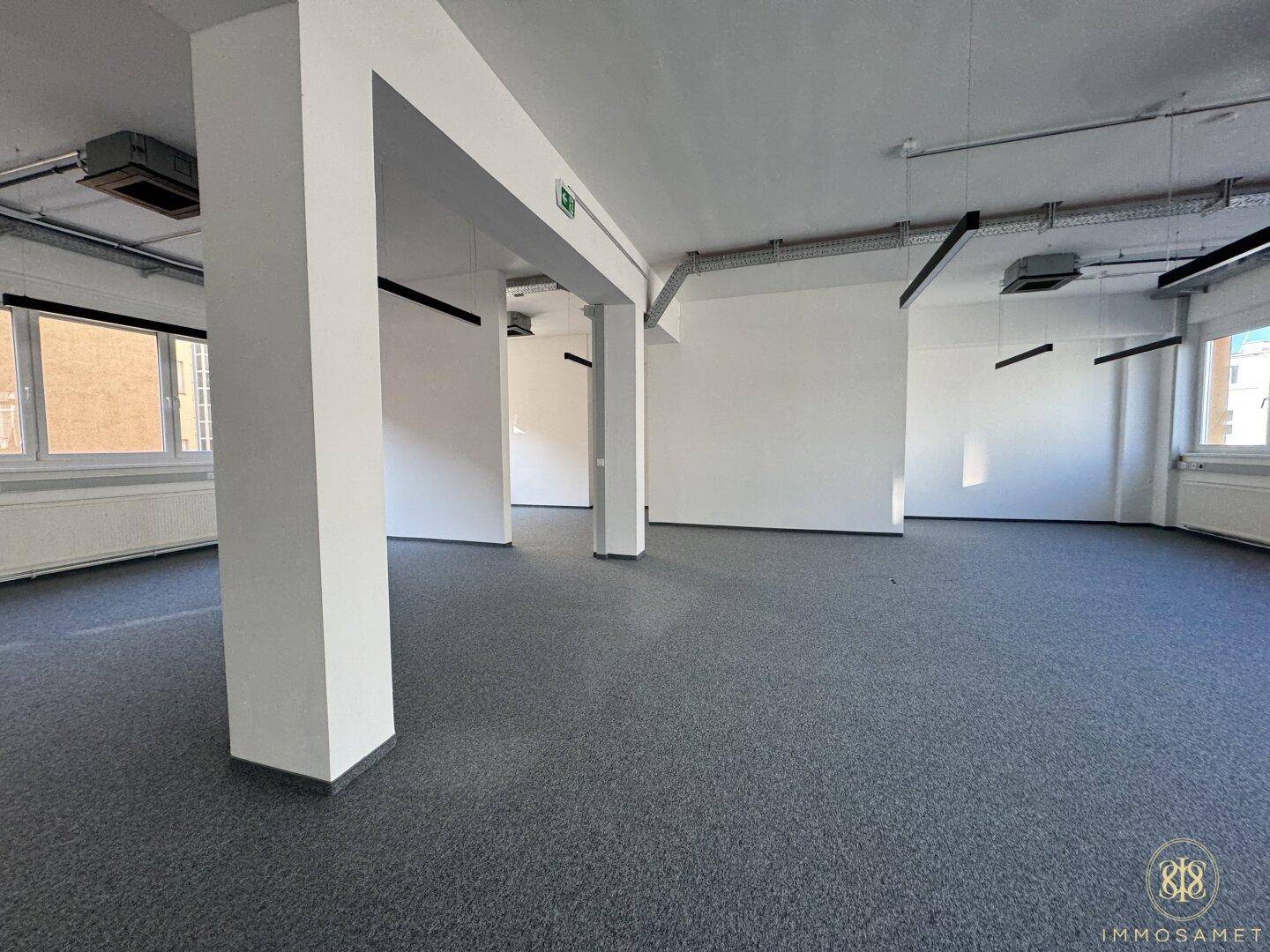 Offener Büroraum mit tragenden Säulen, grauem Teppichboden und industriellem Decken-Design, bietet eine flexible Arbeitsumgebung.