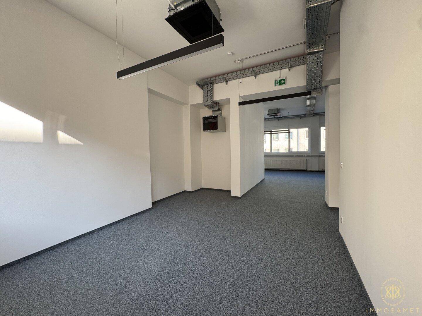 Weitläufiger Büroraum mit grauem Teppichboden, weißen Wänden und sichtbaren Deckeninstallationen, bietet eine funktionale Arbeitsumgebung.