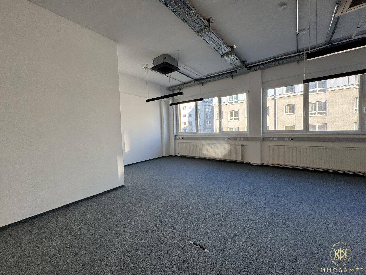 Heller Büroraum mit großen Fenstern, grauem Teppichboden und sichtbaren Deckeninstallationen, bietet eine flexible und moderne Arbeitsumgebung.
