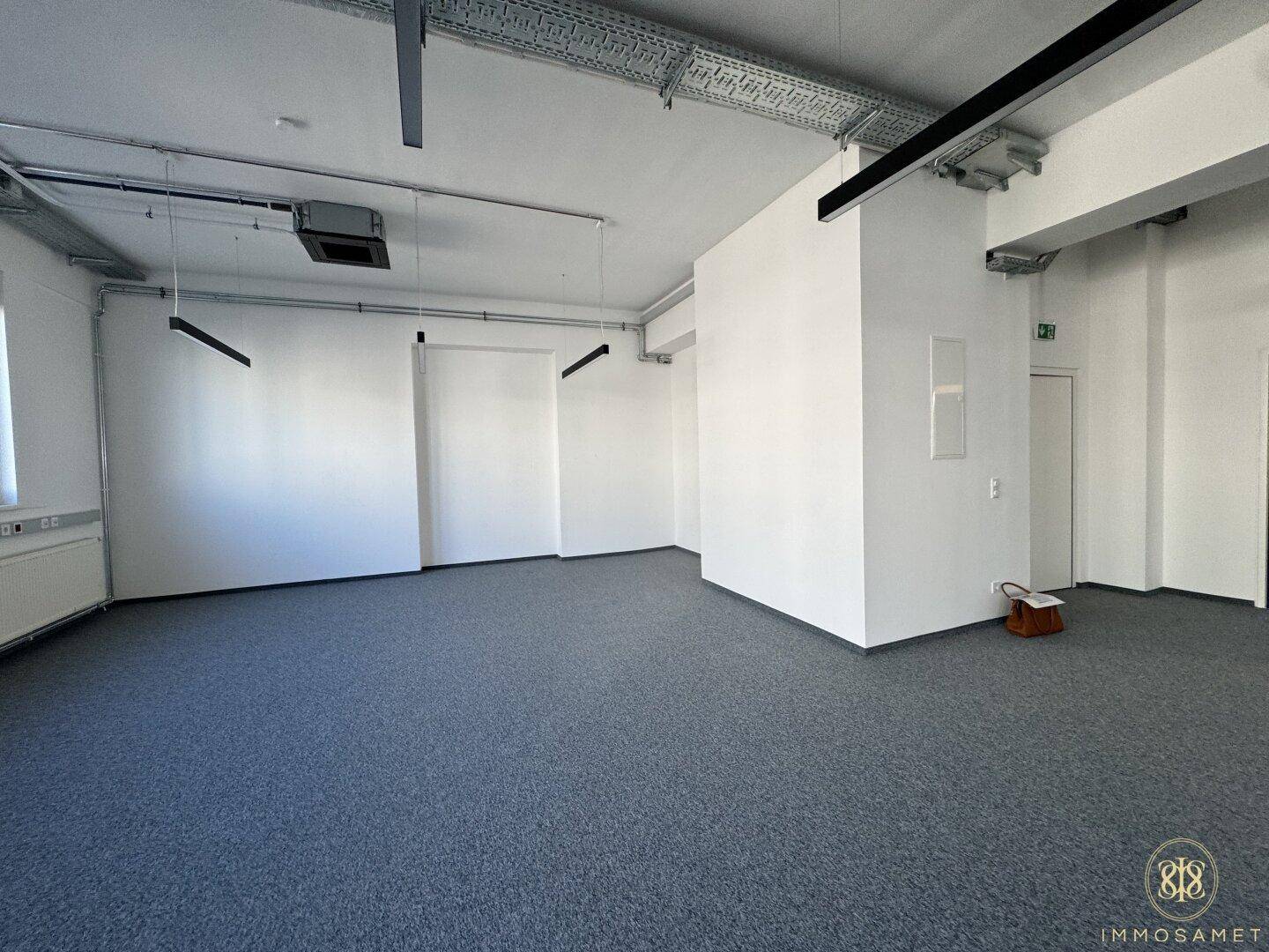 Geräumiger Bürobereich mit grauem Teppichboden, weißen Wänden und sichtbaren Deckeninstallationen, bereit für individuelle Gestaltung.