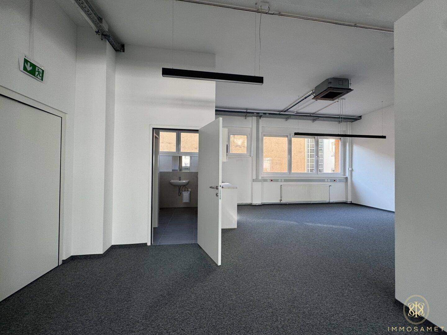 Bürobereich mit grauem Teppichboden und Zugang zu einem Badezimmer, bietet eine praktische und funktionale Raumaufteilung.