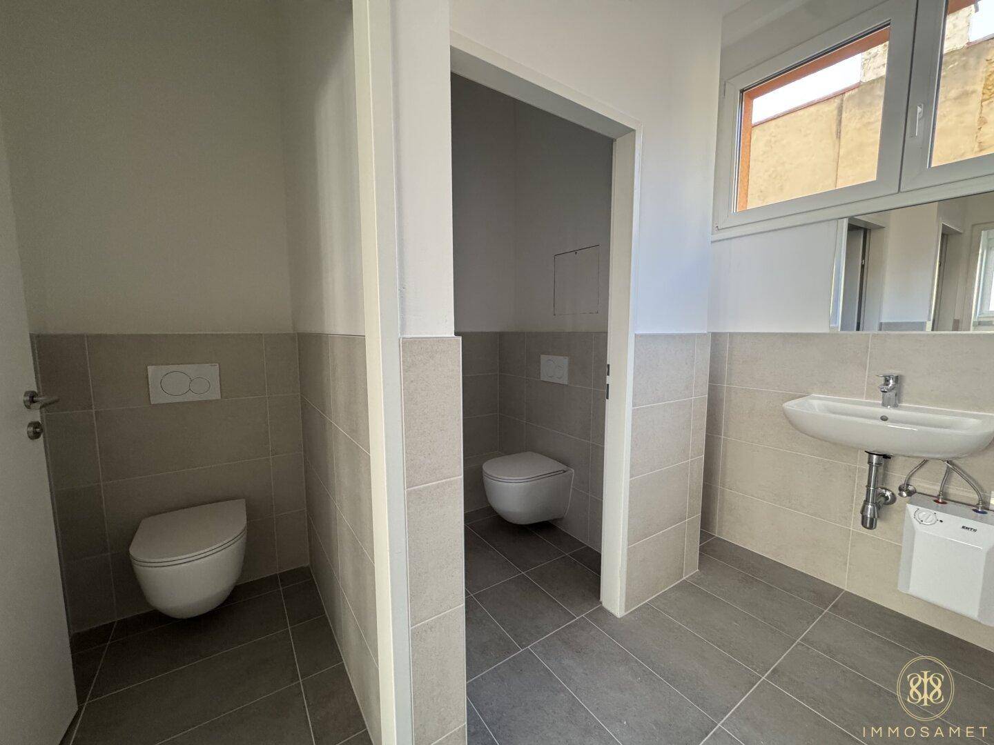 Helles Badezimmer mit zwei wandhängenden Toiletten, einem Waschbecken und modernen Fliesen an Wänden und Boden.