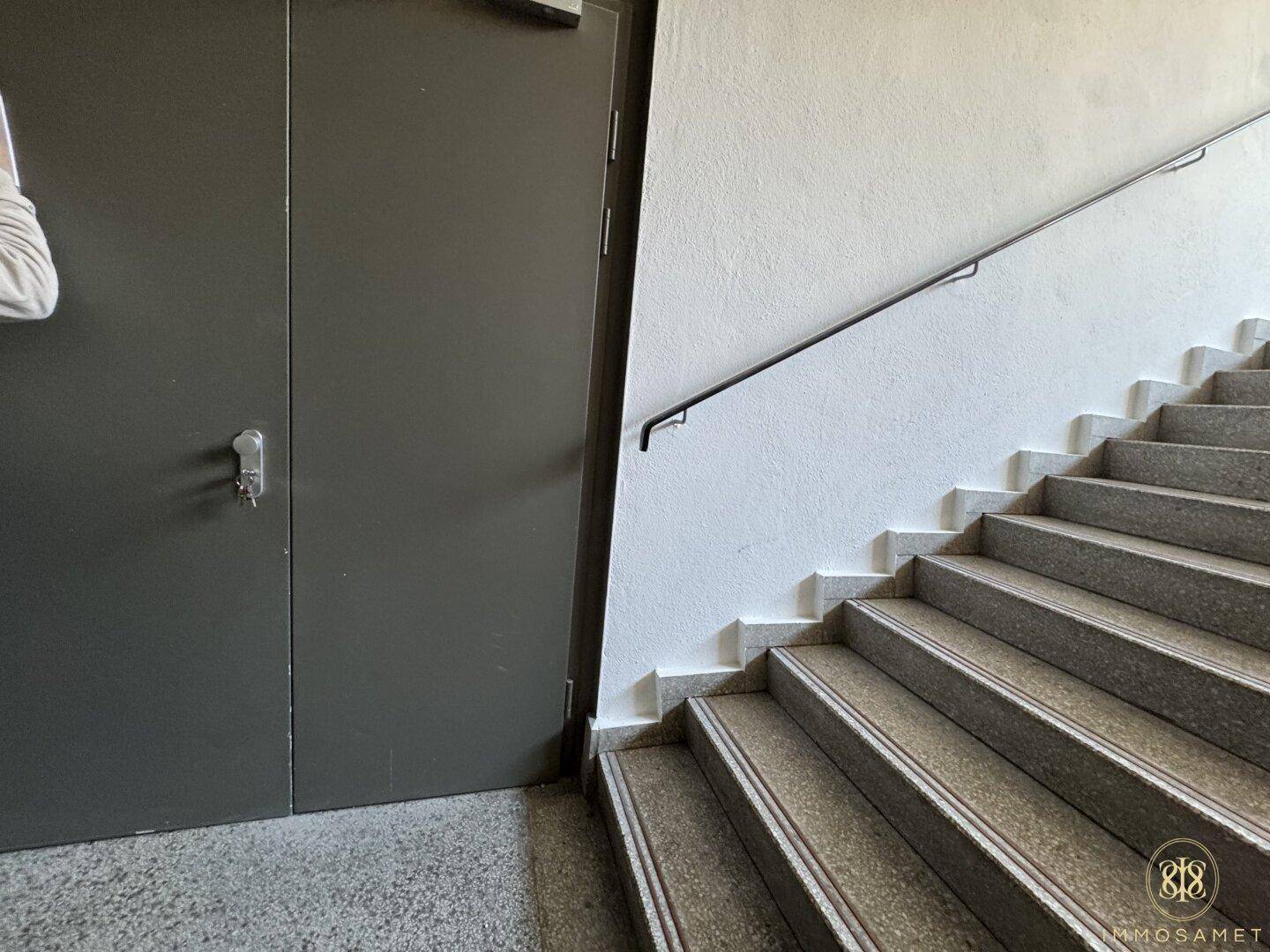 Detailansicht eines Treppenhauses mit Steinstufen und einem einfachen Handlauf an der weißen Wand.