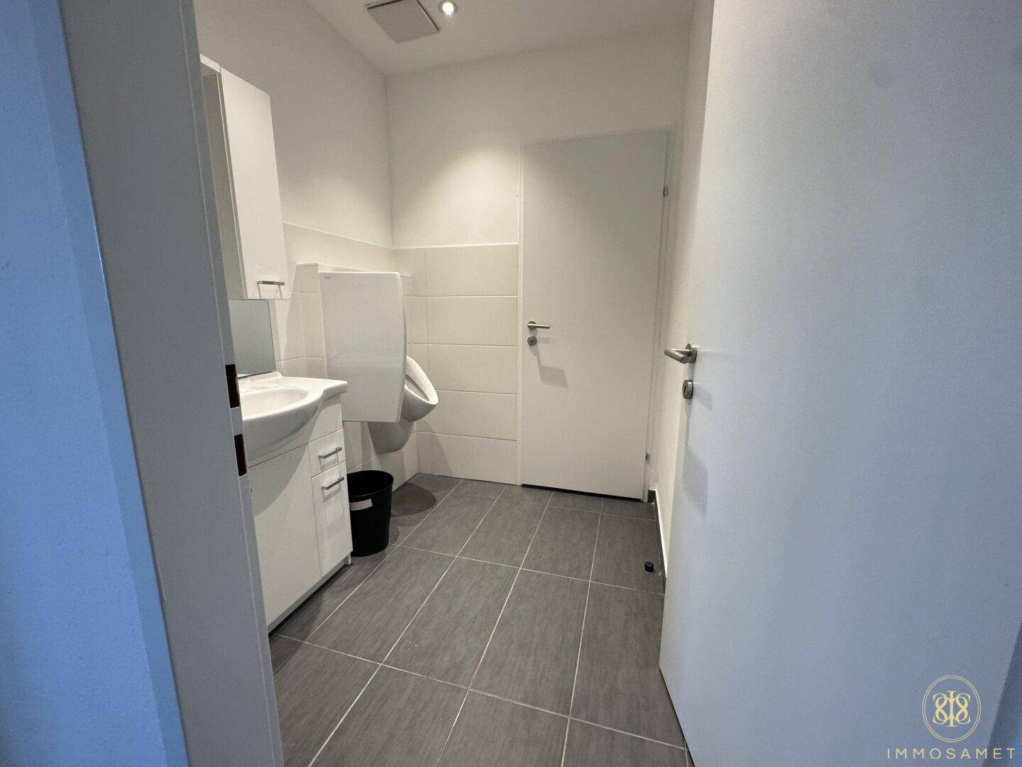 Saubere Toilette mit Fliesenboden, Waschbecken, Wand-WC und Urinal, funktional und hell.