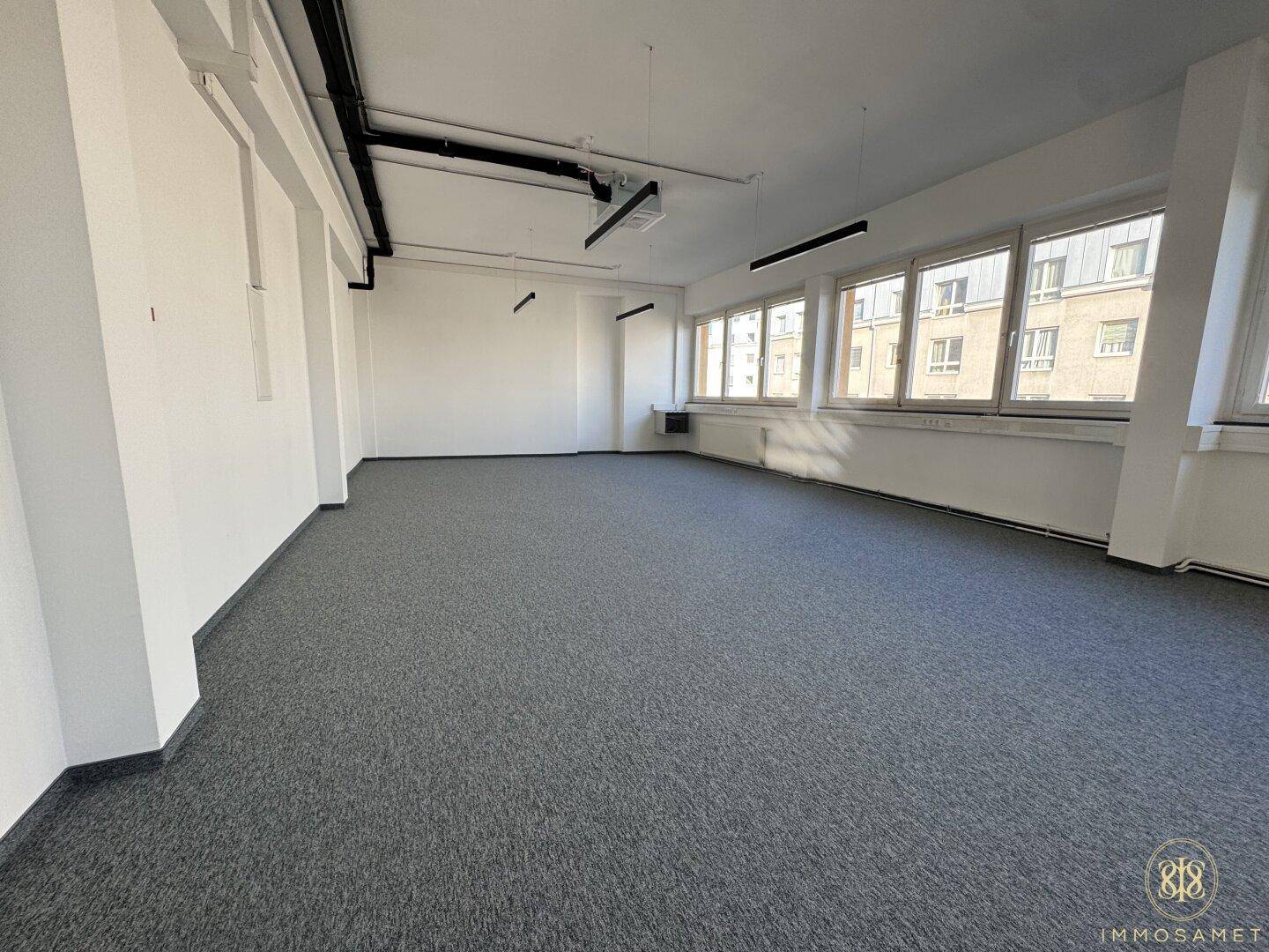 Leerer Büroraum mit Teppichboden und mehreren Fenstern, die für eine helle und offene Atmosphäre sorgen.
