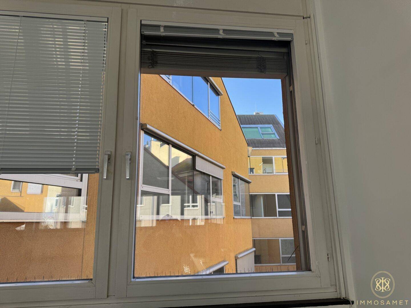 Fenster mit Jalousien und Blick auf die gegenüberliegende Gebäudefassade, die viel Licht reflektiert.