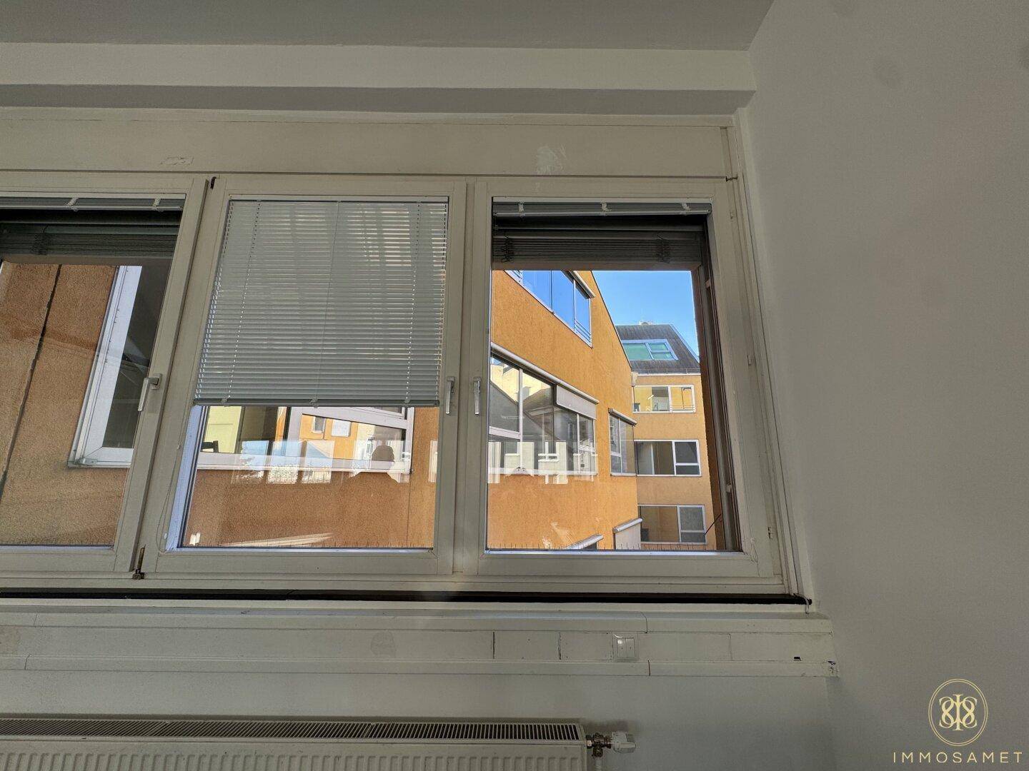 Fenster mit Jalousien und Blick auf die umliegenden Gebäude, die für eine helle Umgebung sorgen.