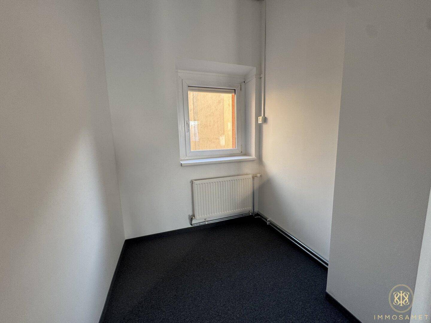 Kleiner, heller Raum mit einem Fenster, Heizkörper und Teppichboden, ideal als Einzelbüro.