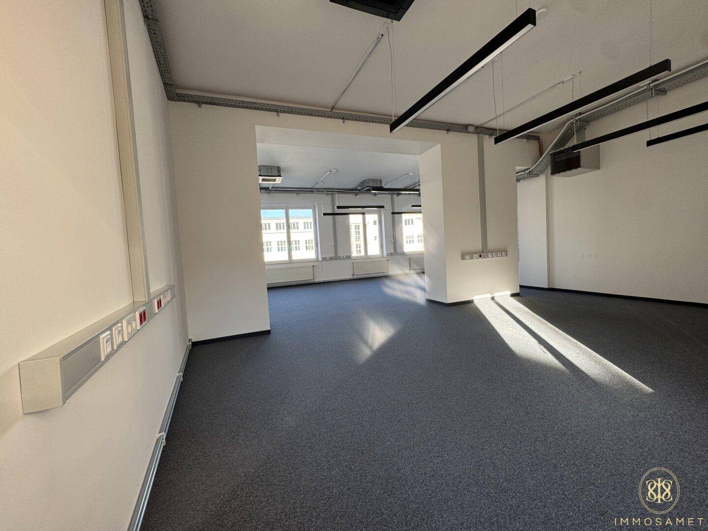 Offener Bürobereich mit Teppichboden, vielen Fenstern und modernen Beleuchtungselementen an der Decke.