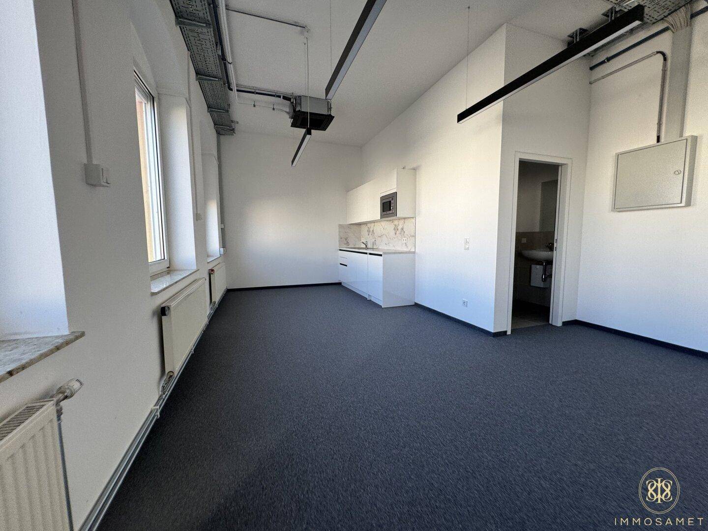 Heller Büroraum mit Teppichboden, integrierter Küchenzeile und Zugang zu einem separaten Toilettenbereich.