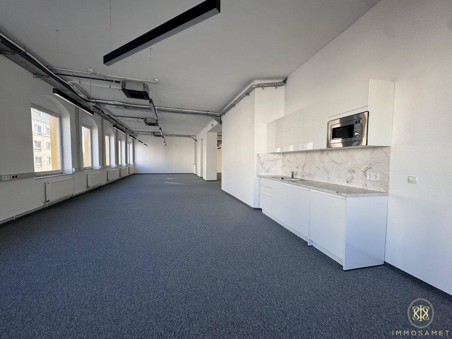 Geräumiges Büro mit Teppichboden, vielen Fenstern und einer modernen, weißen Küchenzeile mit Mikrowelle.