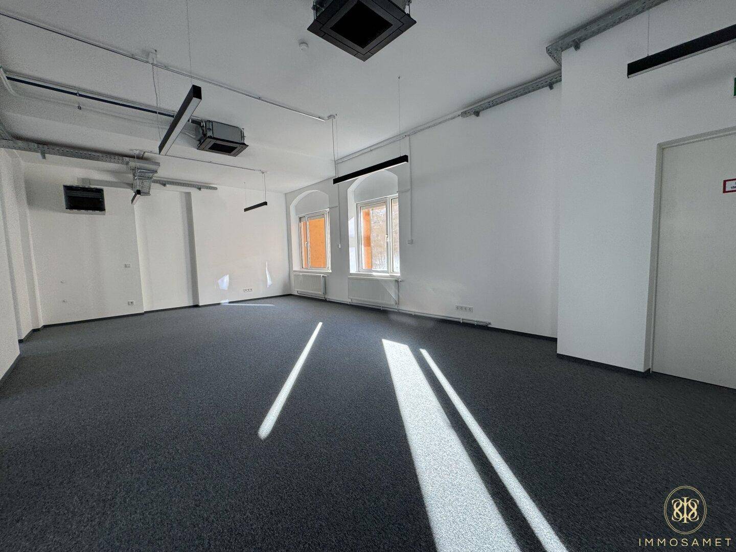 Geräumiger Büroraum mit Teppichboden und mehreren Fenstern, die viel natürliches Licht hereinlassen.