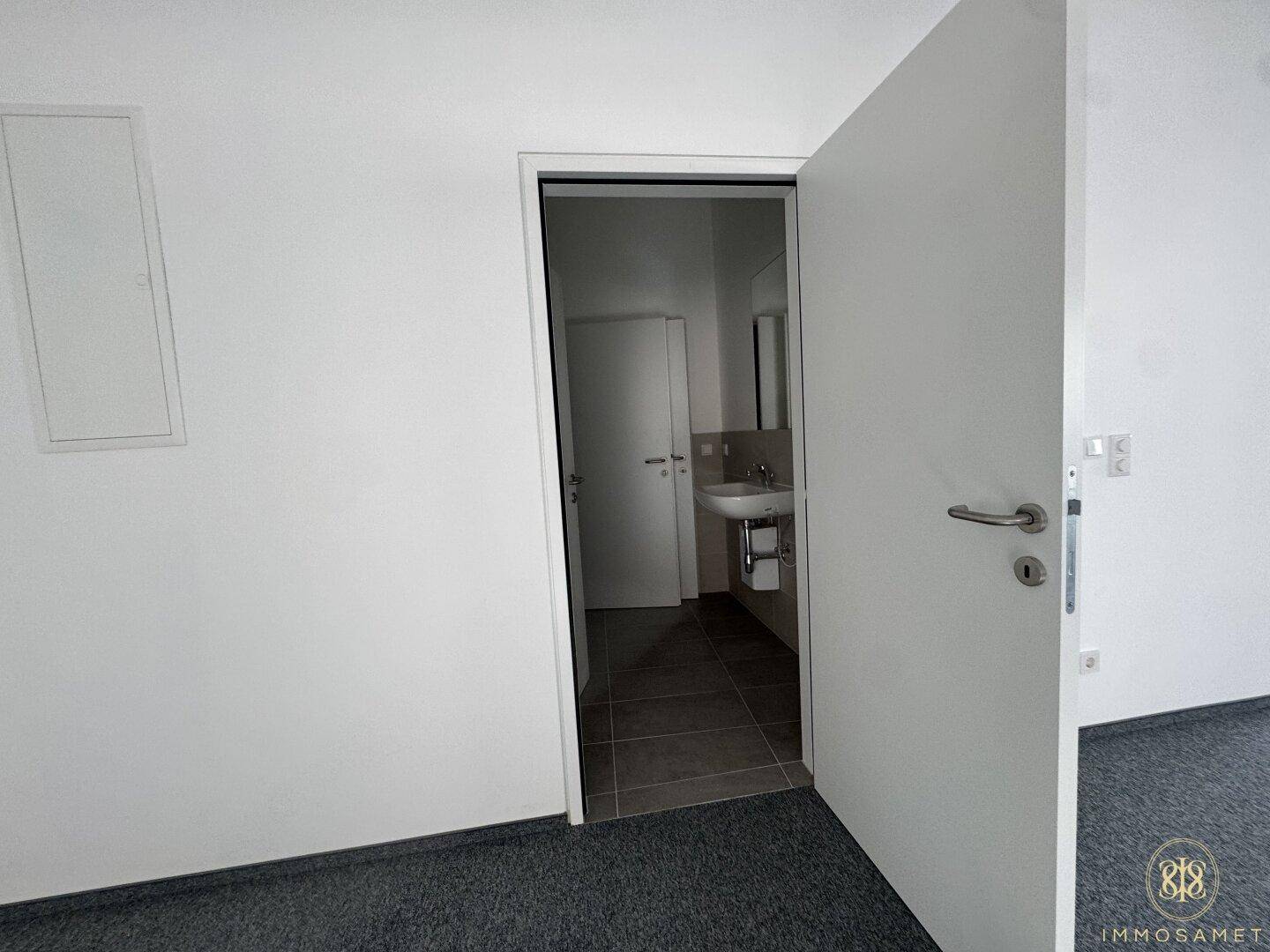 Modernes Badezimmer mit Waschbecken, Spiegel und dunklen Fliesen, zugänglich durch eine offene Tür.