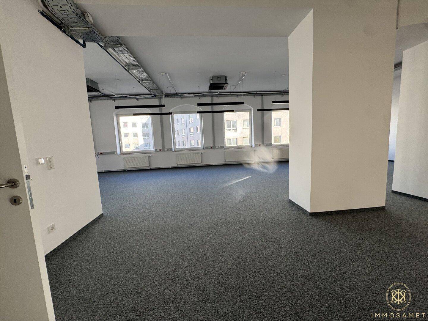 Großer, offener Bürobereich mit Teppichboden, Säulen und einer Reihe von Fenstern, die Tageslicht spenden.