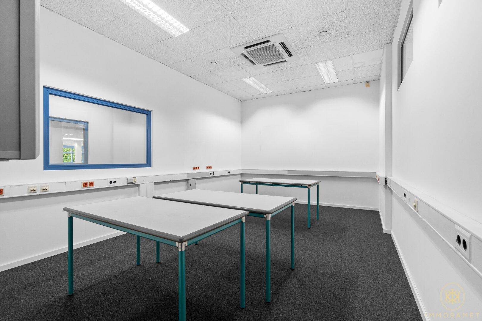 Heller Arbeitsraum mit mehreren Tischen, Teppichboden und blauen Fensterrahmen, ausgestattet für spezielle Tätigkeiten.
