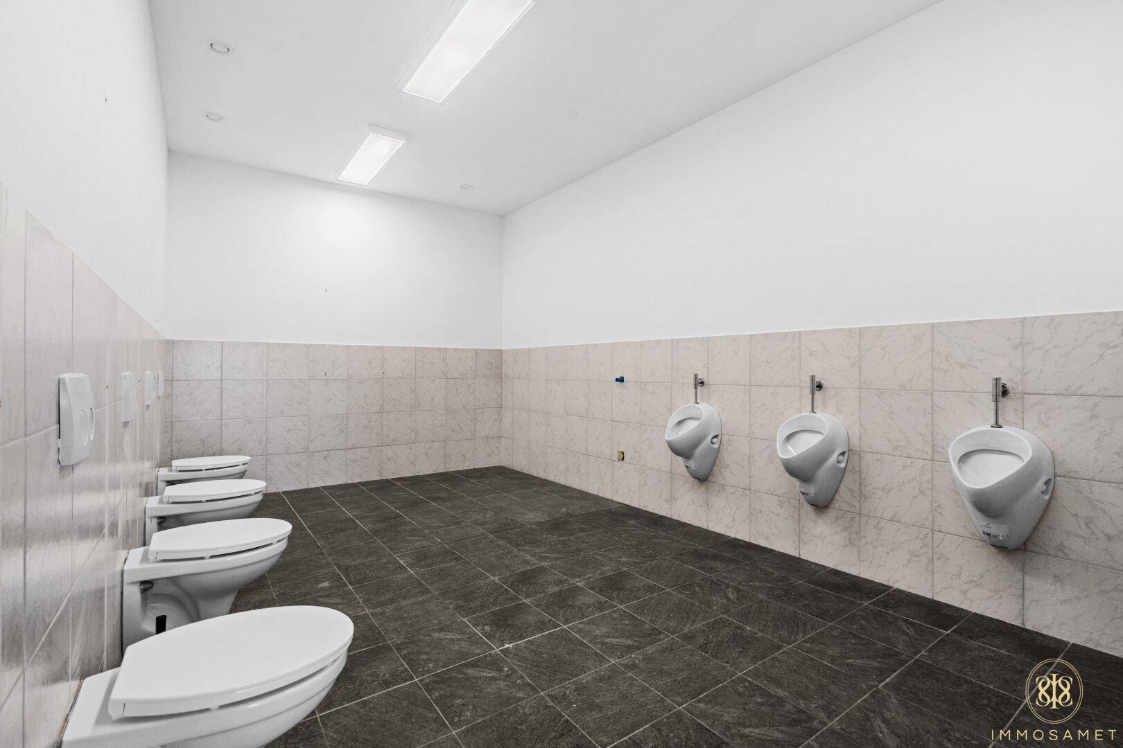 Geräumiger öffentlicher Waschraum mit mehreren Toiletten und Urinalen, ausgestattet mit Fliesenboden und Wandfliesen.