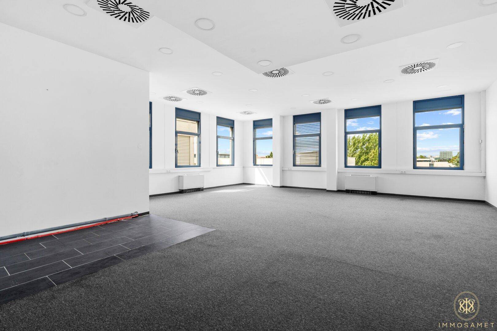 Heller Büroraum mit Teppichboden und Fliesenbereich, ausgestattet mit vielen Fenstern und Lüftungssystemen.