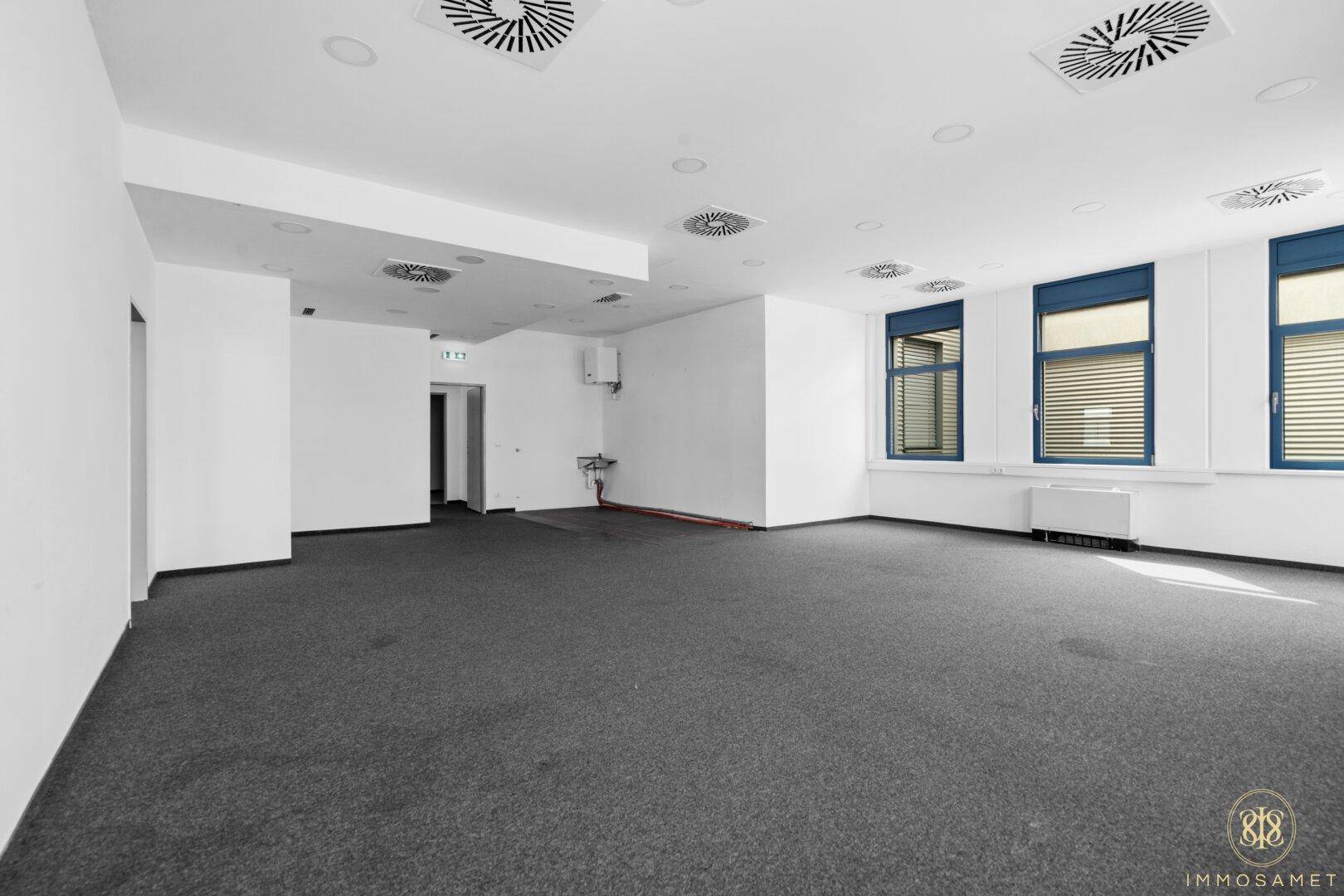 Offener Bürobereich mit Teppichboden und Zugang zu weiteren Räumen, gut beleuchtet durch Fenster.