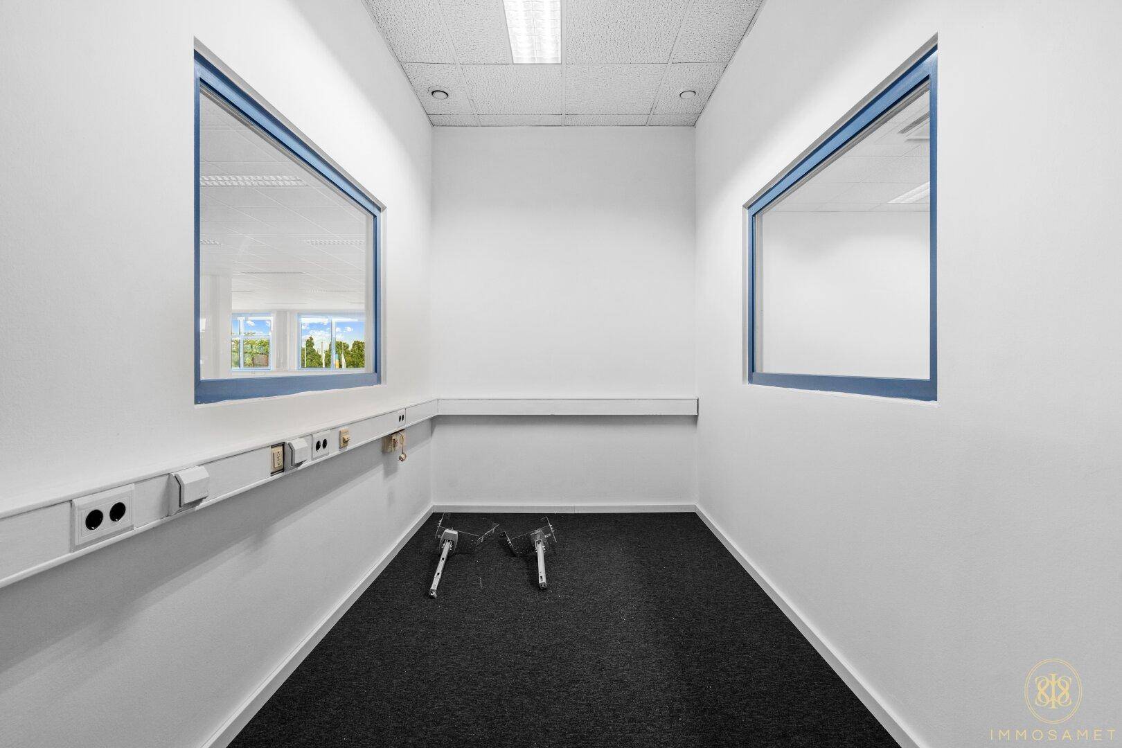 Kleiner, heller Raum mit zwei Fenstern und Teppichboden, geeignet als Einzelbüro oder Abstellraum im Gebäude.