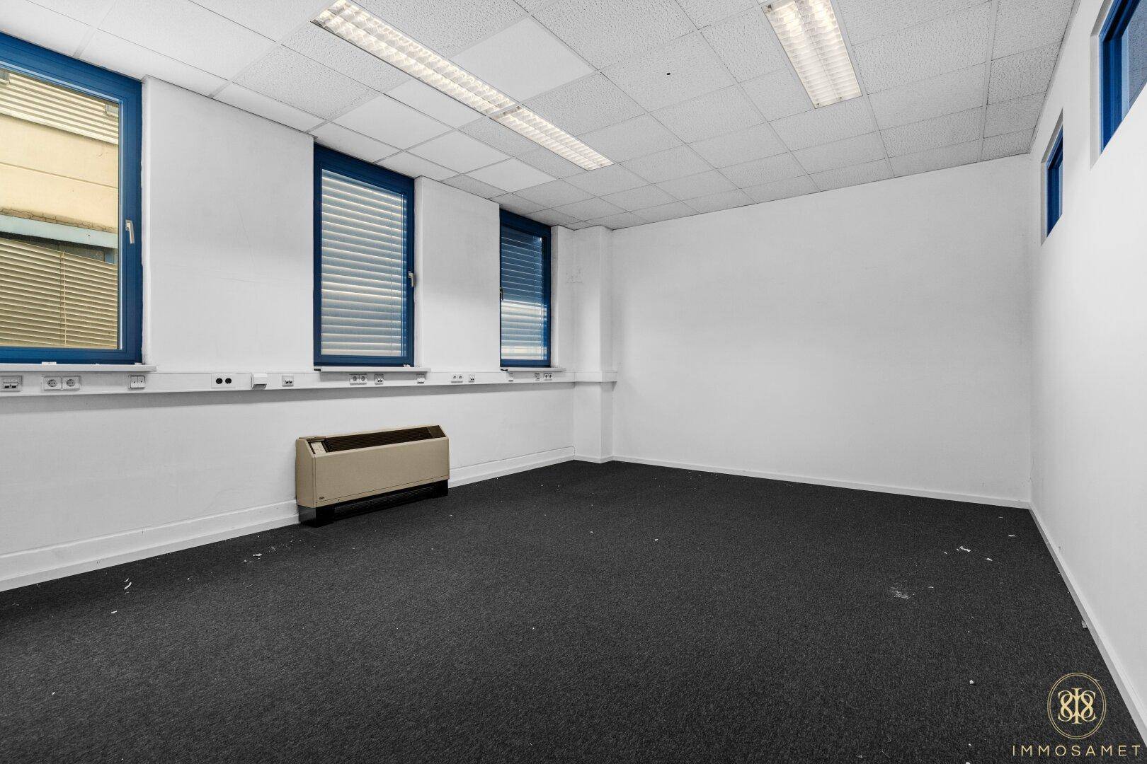 Leerer Büroraum mit Teppichboden, blauen Fensterrahmen und Heizkörper, bereit zur Einrichtung und Nutzung.