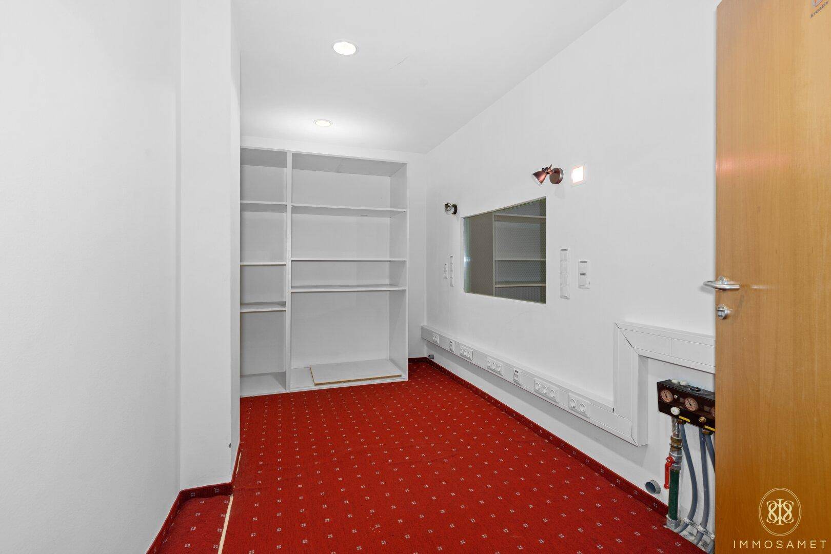 Kleiner Büroraum mit rotem Teppichboden und eingebauten Regalen.