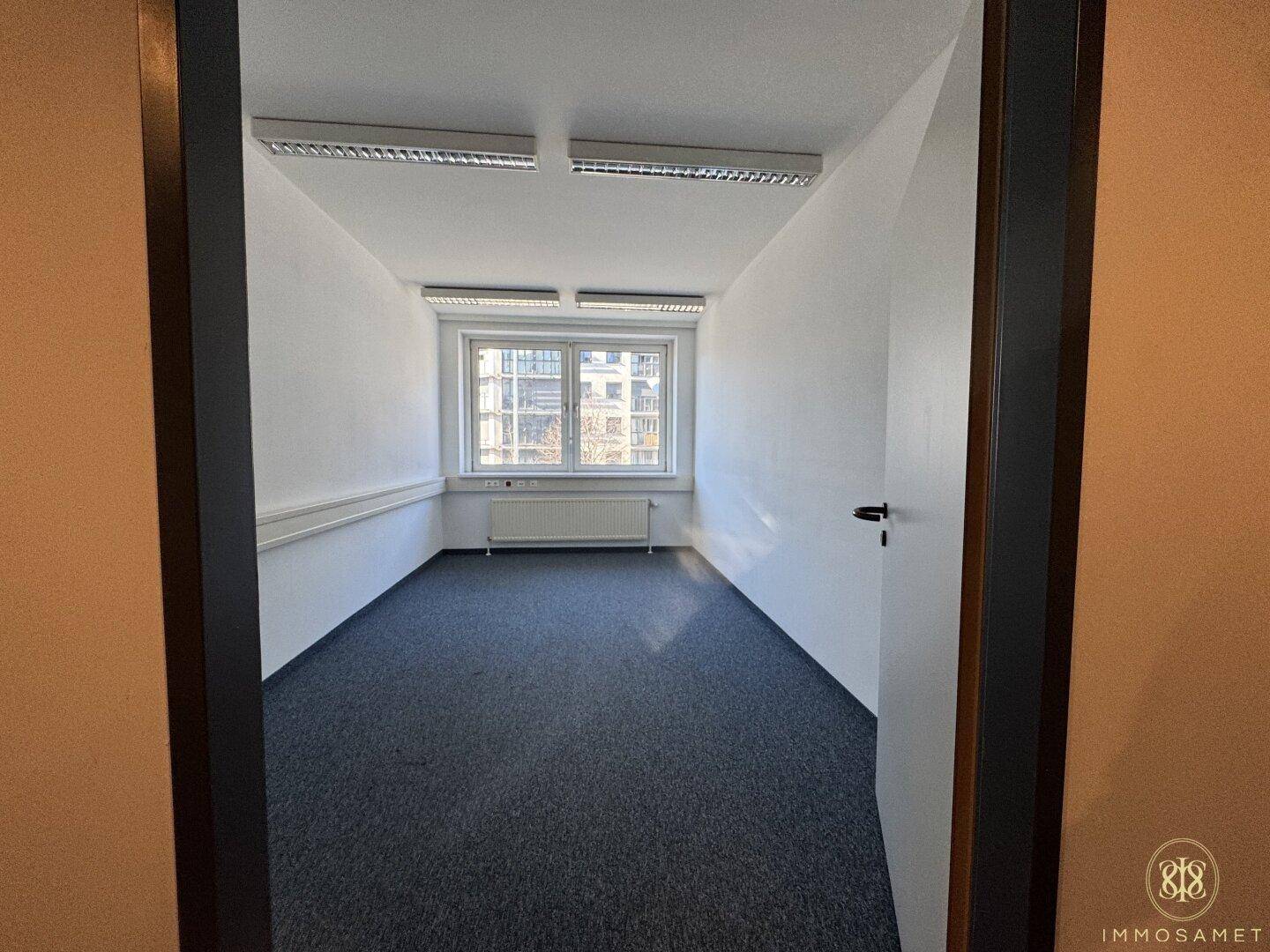 Blick durch eine Tür in ein leeres Büro mit Teppichboden und einem großen Fenster am Ende des Raumes.