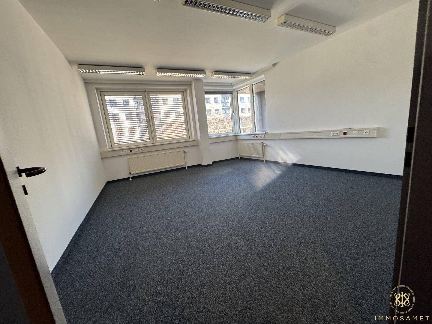 Helles Büro mit grauem Teppichboden und einer breiten Fensterfront, die den Raum mit Tageslicht füllt.