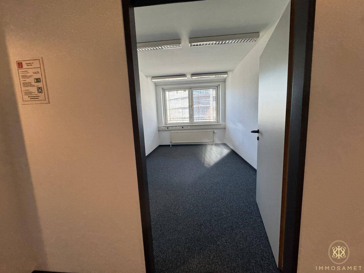 Leeres Büro mit Teppichboden und einem Fenster, das den Raum mit natürlichem Licht erhellt.