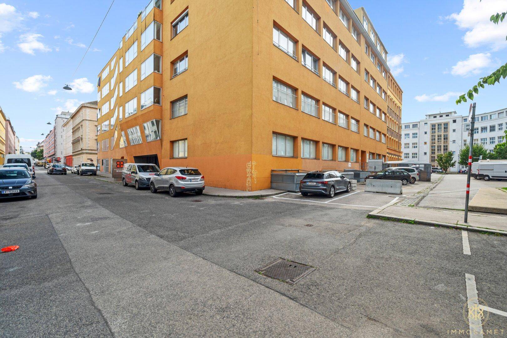 Ansicht eines modernen orangefarbenen Gebäudes an einer Stadtstraße mit geparkten Autos und klarem Himmel.