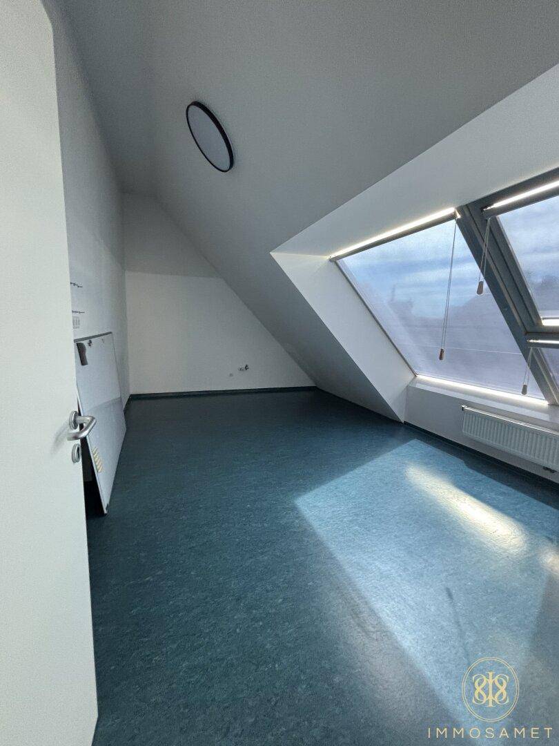 Heller Dachgeschossraum mit schräger Decke, großem Fenster und dunklem Bodenbelag, bietet viel Tageslicht.