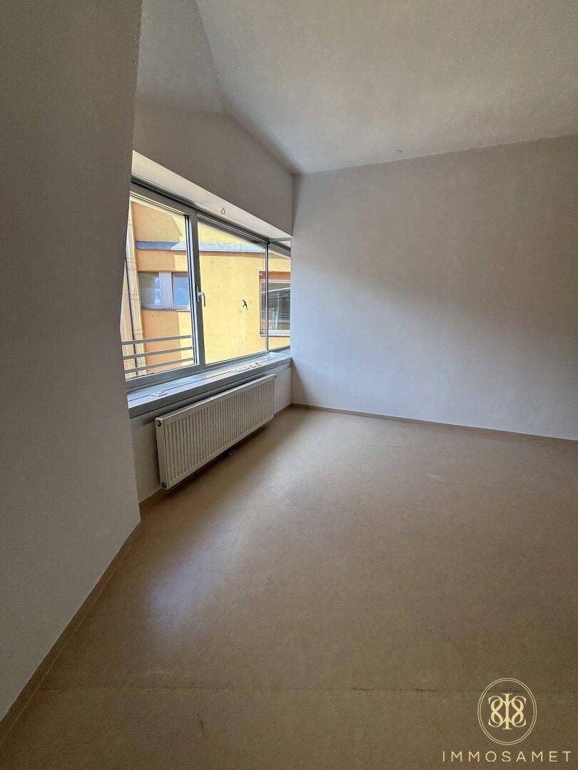 Heller Raum mit beigem Bodenbelag, weißen Wänden und einem Fenster mit Blick auf umliegende Gebäude.