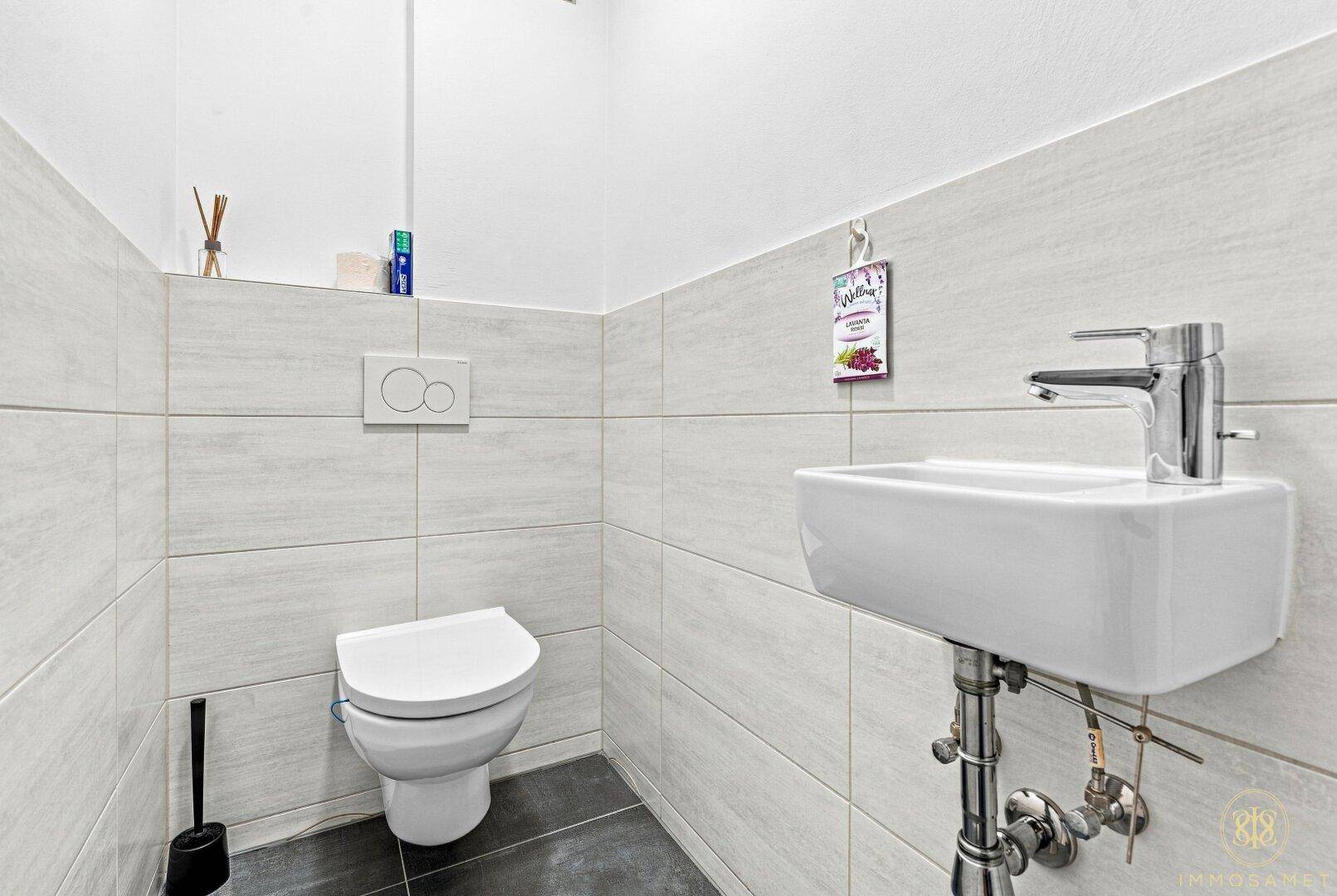 Modernes Gäste-WC mit Wand-WC, kleinem Waschbecken und hellen Fliesen an den Wänden.