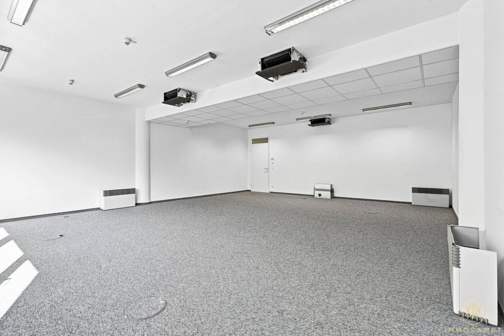Heller Büroraum mit Teppichboden, weißen Wänden, abgehängter Decke und sichtbaren Klimaanlagen.