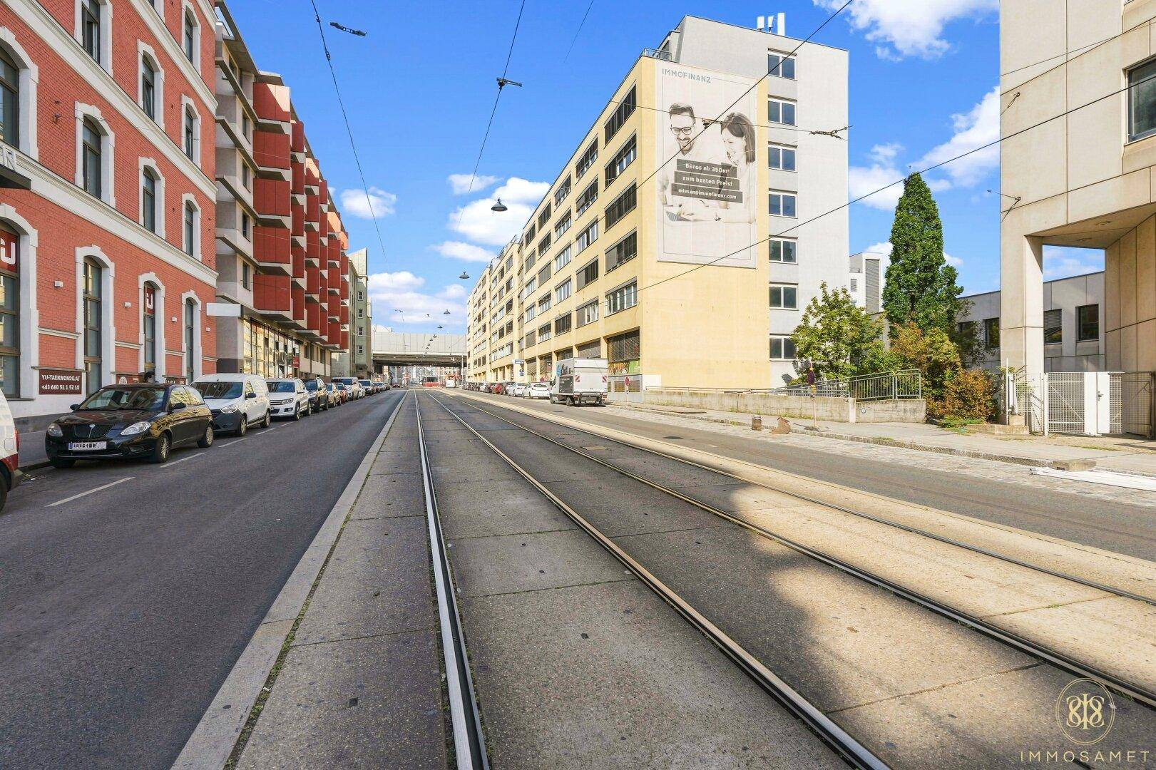 Straßenansicht mit Straßenbahnschienen, die ein langes, gelbes Gebäude und weitere Gebäude in der Ferne zeigt.
