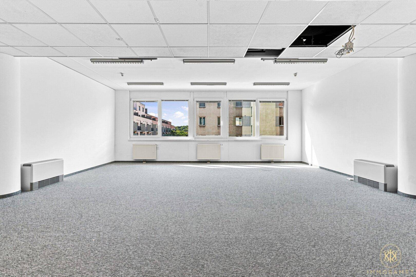 Heller Büroraum mit großen Fenstern, die einen Blick auf umliegende Gebäude bieten.