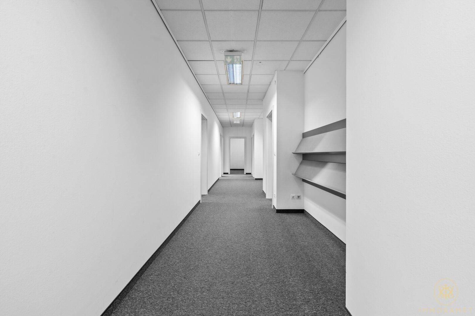 Langer, heller Bürogang mit Teppichboden und Deckenbeleuchtung, der zu weiteren Räumen führt.