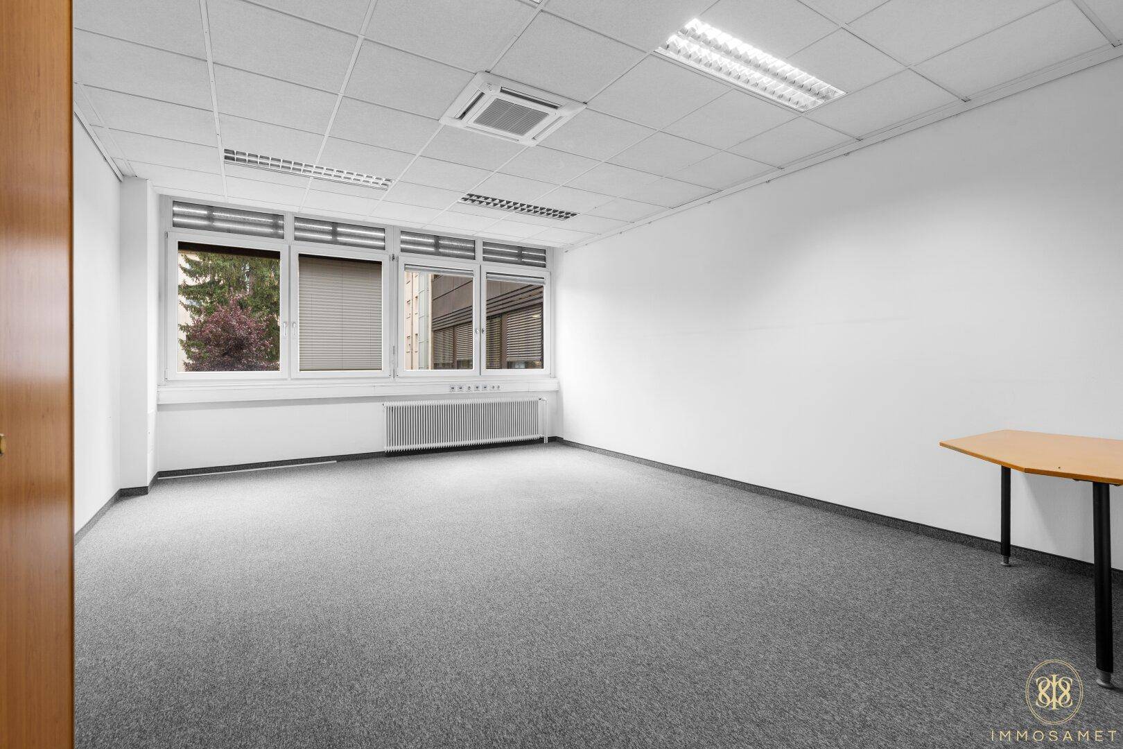 Leeres Büro mit Teppichboden, großen Fenstern und einem kleinen Tisch, bereit zur individuellen Gestaltung.