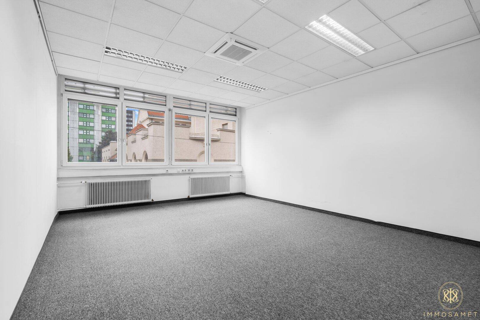 Helles Büro mit Teppichboden und Fenstern, die einen Blick auf die umliegende Stadtbebauung ermöglichen.