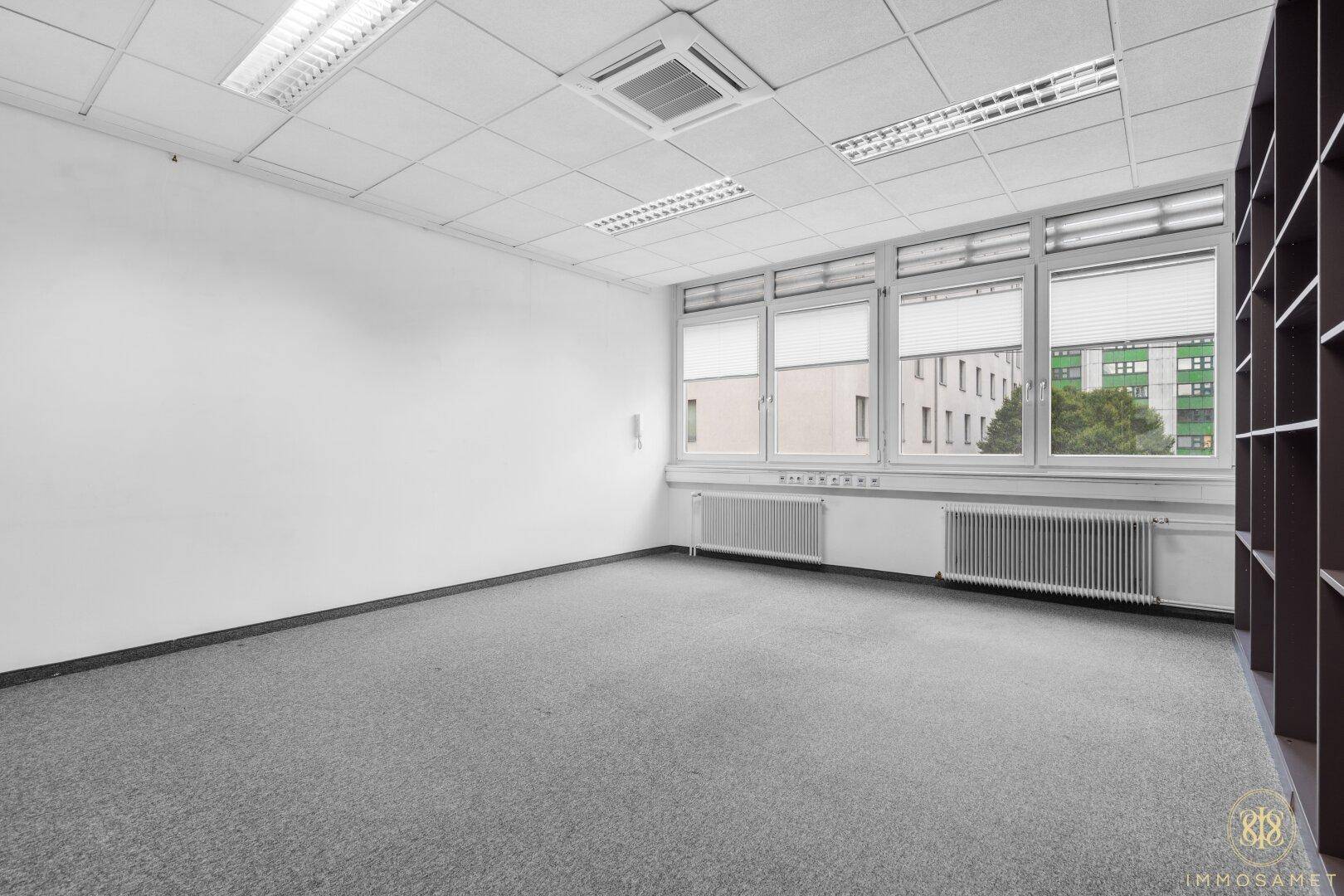 Leeres Büro mit Teppichboden, Fenstern und einem praktischen Einbauregal für Stauraum.