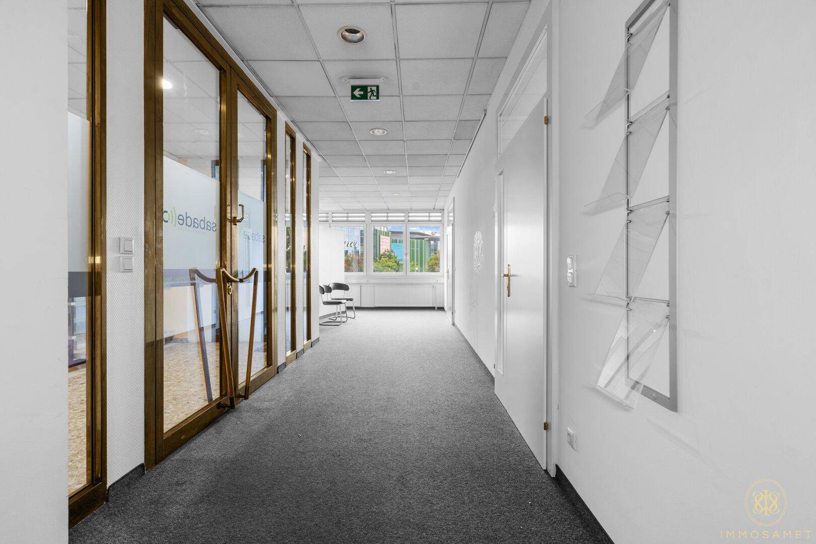Büroflur mit Glastüren zu den Büros, Teppichboden und einem gut sichtbaren Notausgangsschild.