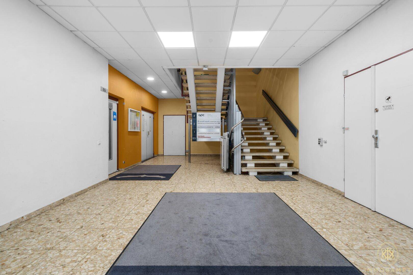 Eingangsbereich des Gebäudes mit Treppe, Aufzug und gefliestem Boden, der zu den oberen Etagen führt.