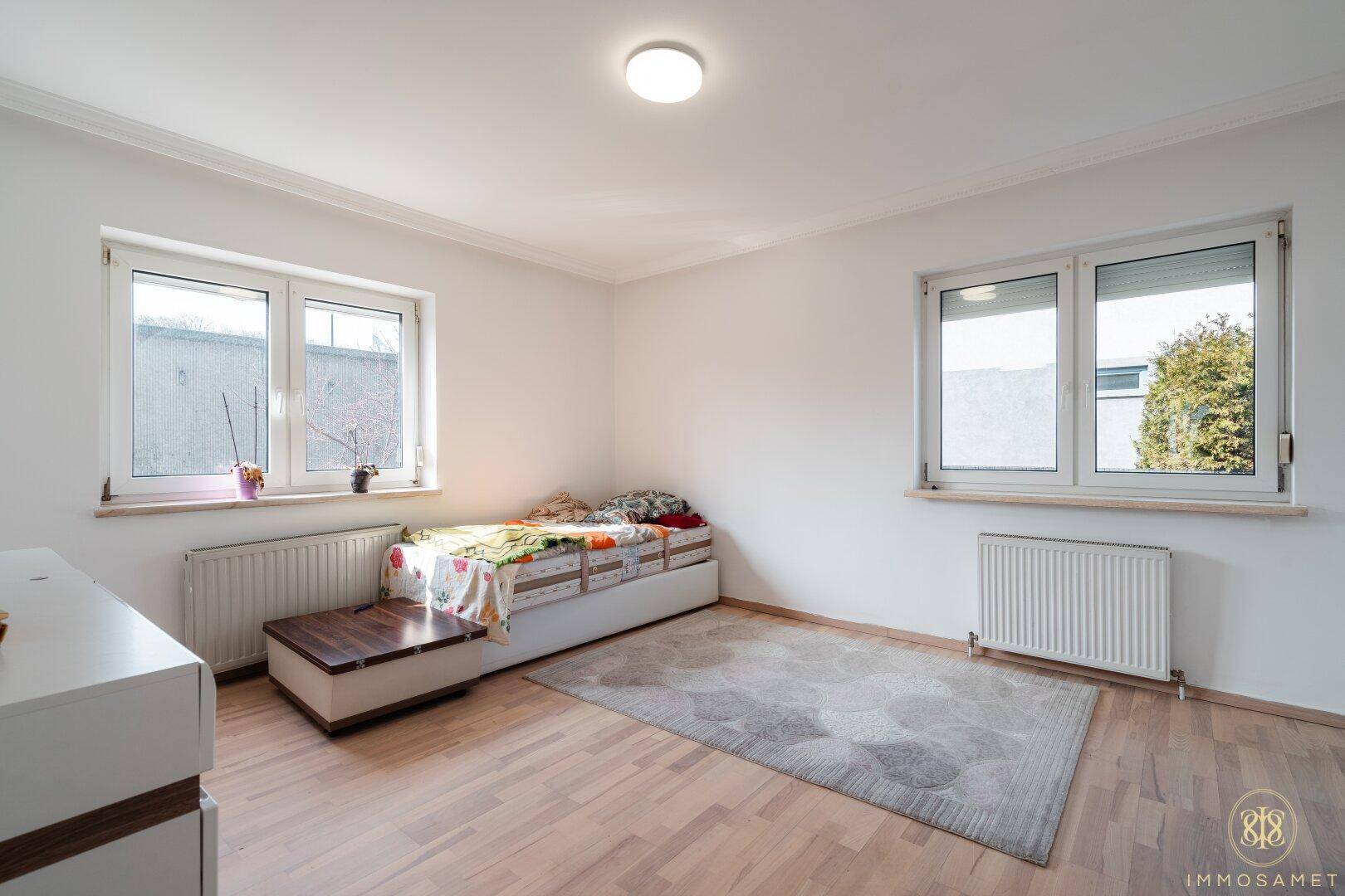 Helles Schlafzimmer mit Holzboden, weißen Wänden, Einzelbett und zwei großen Fenstern für Tageslicht.