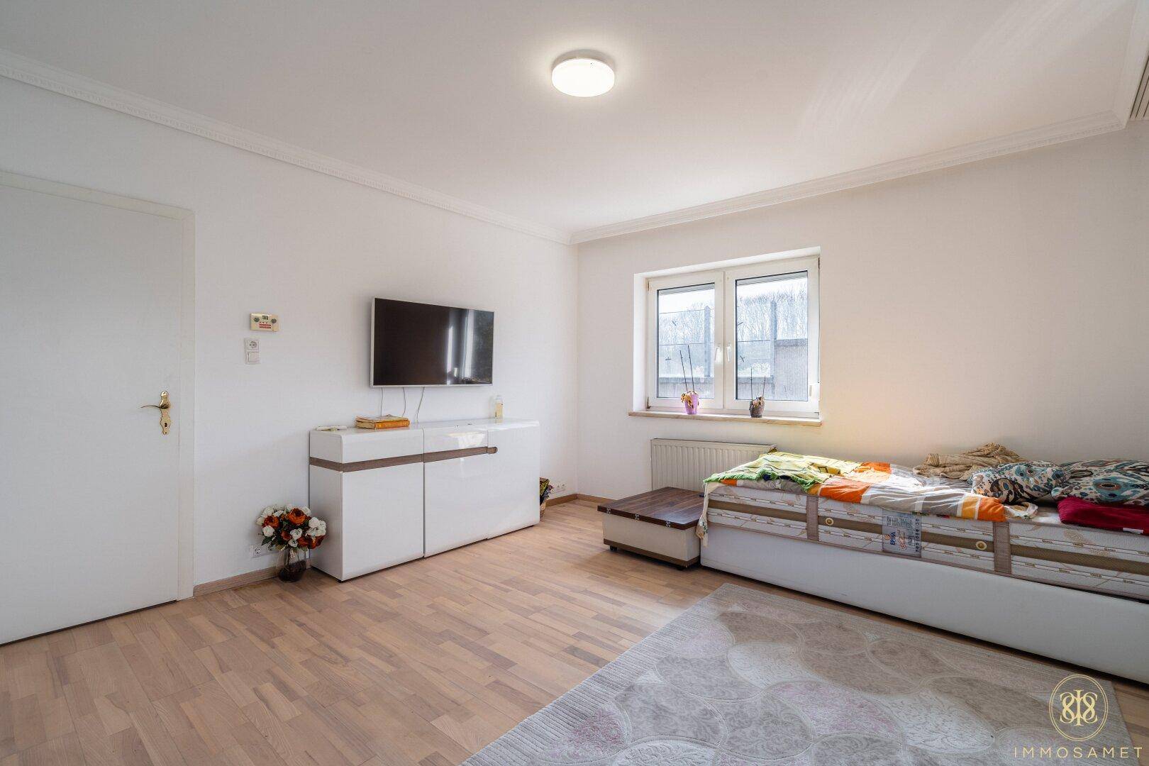 Schlafzimmer mit Holzboden, modernem TV-Möbel und viel Tageslicht durch das große Fenster.