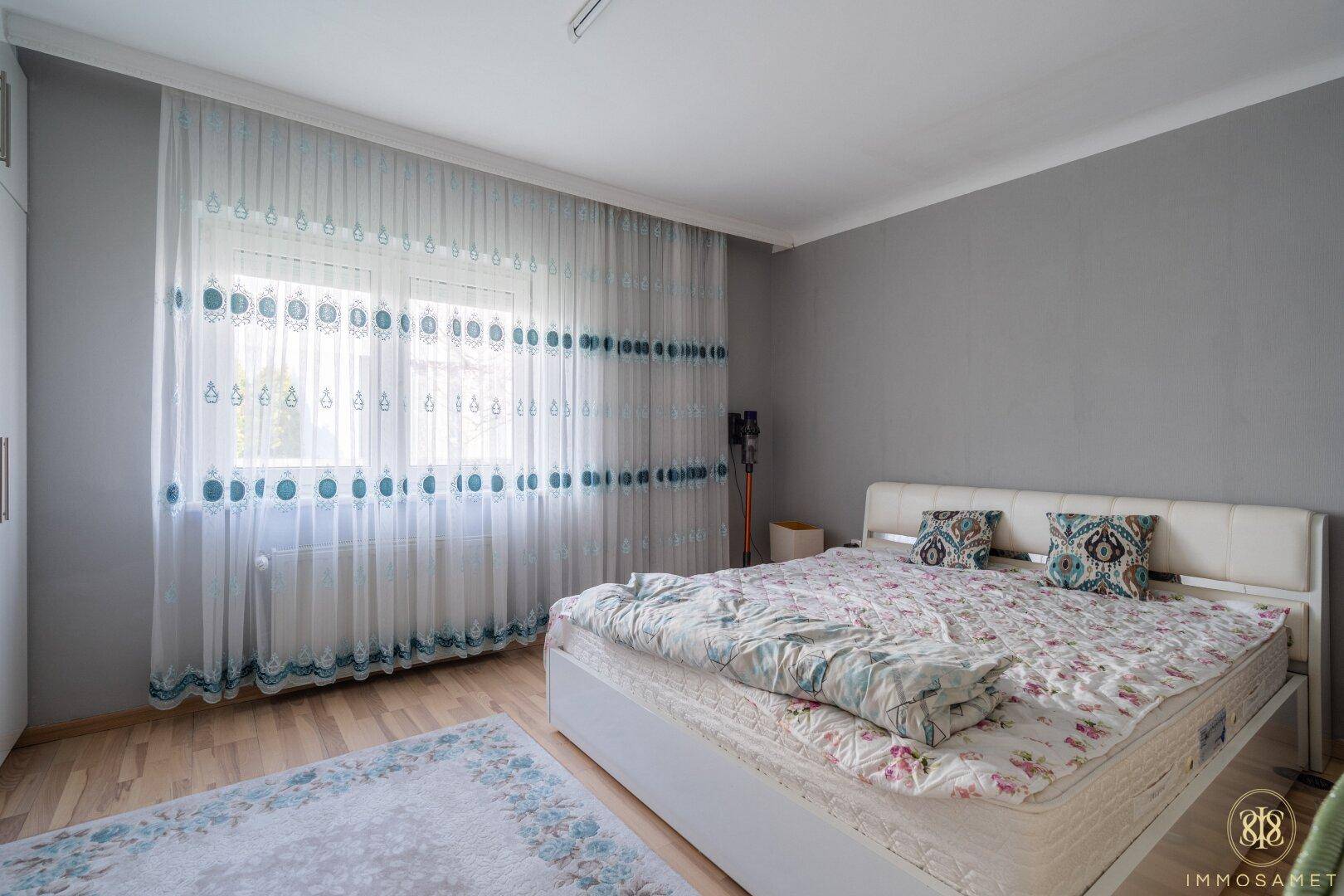 Geräumiges Schlafzimmer mit grauen Wänden, Holzboden und großem Fenster mit hellen Vorhängen.