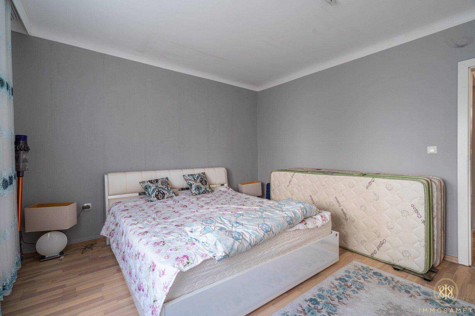 Schlafzimmer mit Doppelbett, grauen Wänden und Holzboden, bietet Platz für zusätzliche Möbel.