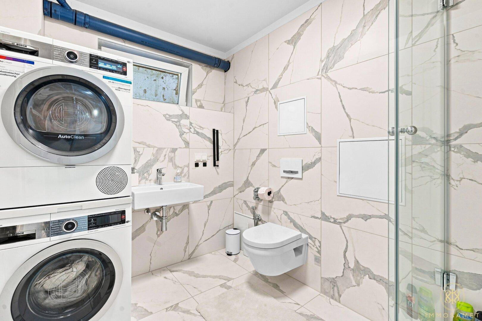 Modernes Badezimmer mit Waschmaschine, Trockner, WC, Waschbecken und Duschbereich in Marmoroptik.